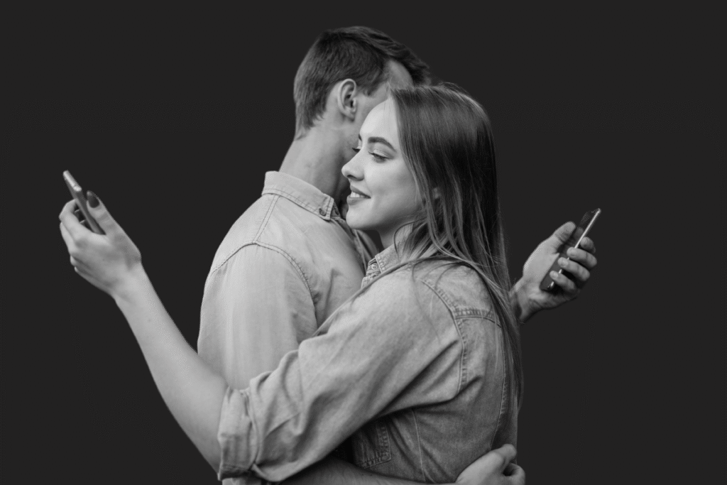 Un ragazzo ed una ragazza si abbracciano, ma guardano i loro cellulari. 